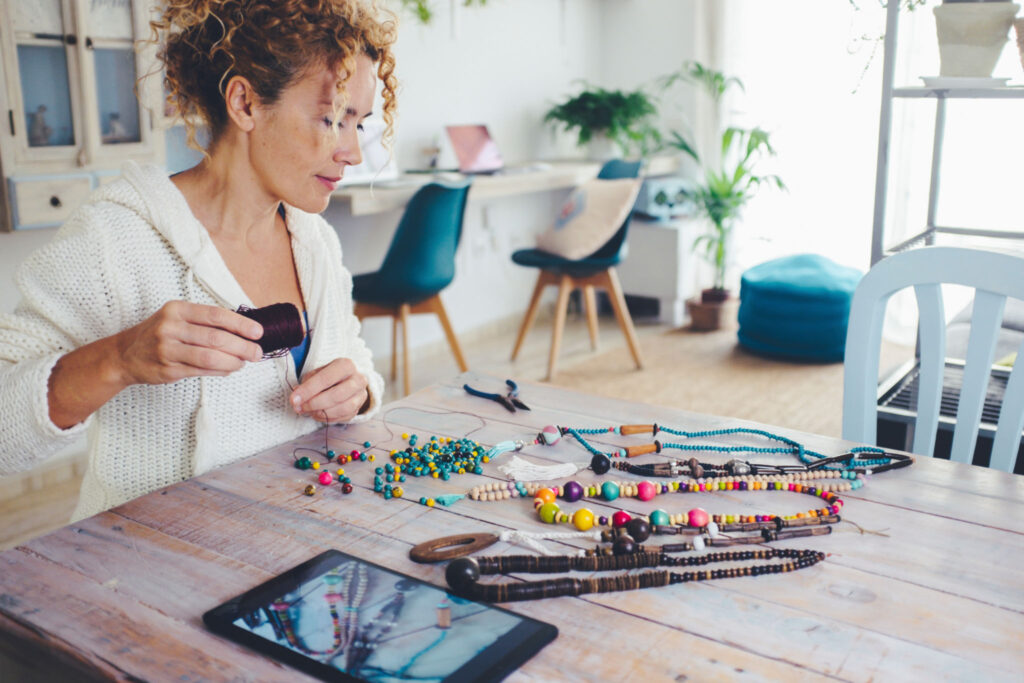 People in indoor leisure activity concept lifestyle. One adult woman doing beads necklace on the table in living room with tablet device on. Modern alternative new business work online selling on web