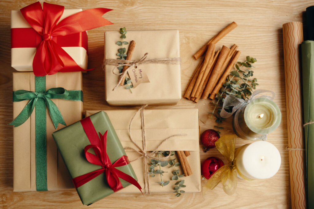 Various gift boxes wrapped in paper and decorated with ribbons and sprigs, accompanied by cinnamon sticks and candles, arranged on wooden table surface