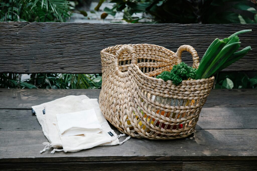 Wicker basket with heaped organic vegetables placed on wooden bench near reusable grocery bags in lush garden